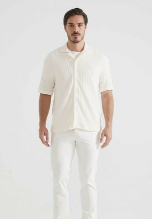 Homme debout, regardant vers l'avant, portant une chemise crème à manches courtes texturée et un pantalon blanc, sur fond blanc uni.