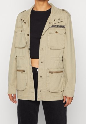 Khaki utilityjack biedt meerdere zakken, accenten van zilverkleurig hardwaren en een gemêleerde strip op de borst. Gemaakt van duurzaam fabric.