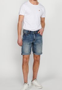 White short-sleeve t-shirt with a small embroidered detail, paired with rolled-up blue denim shorts and white sneakers. Casual outfit.