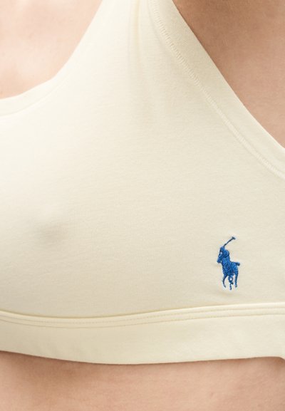 Κοντινό πλάνο στήθους που φοράει σουτιέν εκρού χρώματος με μικρό μπλε λογότυπο παίκτη πόλο κεντημένο στο ύφασμα.