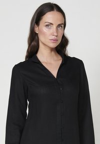 Cămașă neagră cu mâneci lungi, cu un guler și un sistem de închidere cu butoni, confecționată dintr-un material texturat, cu o croială ajustată. Nu sunt vizibile modele sau detalii metalice distincte.