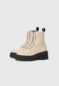 Beige snørebukser med en kraftig svart såle og trekkfliker bak, med metalliske maljer og en jevn overflate.