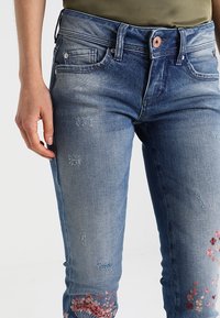 Blue denim jeans with a fitted design, featuring distressed detailing and floral embroidery in pinks at the lower leg. Smooth texture.
