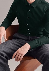 Mężczyzna w ciemnozielonej koszuli zapinanej na guziki i szarych spodniach siedzi na drewnianym krześle, z rękami spoczywającymi na udach.