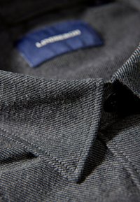 Donker denim overhemd met een gestructureerd oppervlak, voorzien van een blauwe label en een zwarte knop. Opvallende kraag en stikseldetails.