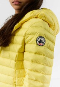 Gele opblaasbare jas met capuchon, voorzien van een horizontaal doorgestikt ontwerp en een ronde logopatch op de mouw. Gladde textuur.