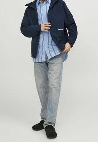 Giacca impermeabile blu navy con cappuccio, abbinata a una camicia a righe azzurro chiaro con bottoni e jeans grigio chiaro. Scarpe nere.