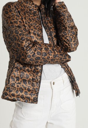 Kvinde iført en brun og sort leopardmønstret quiltet jakke over en grå skjorte og hvide bukser, med armene krydset.