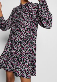 Robe noire à fleurs avec manches longues. Présente des motifs floraux colorés en rose, violet et vert sur un design fluide à volants.