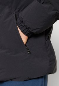 Hand delvis inne i den dragkedjade fickan på en svart jacka med elastiska ärmmuddar, blå byxor synliga nedanför.