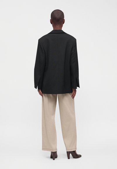 Personne aux cheveux courts portant un blazer noir ample, un pantalon beige à jambes larges et des chaussures à talons foncées, debout de dos sur un fond blanc.