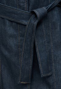 Ζώνη από σκούρο μπλε τζιν ύφασμα δεμένη σε έναν απλό κόμπο γύρω από ενδύματα με ορατή πορτοκαλί ραφή.