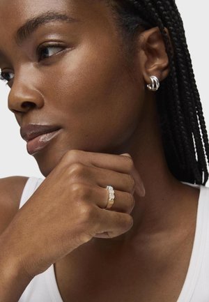 Gold ring with three gemstones, including a central light blue stone, worn on a hand resting against a chin. Model with braided hair and silver earrings.