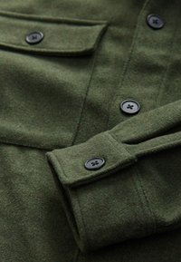 Gros plan sur la manche d'une veste en laine vert foncé avec des boutons noirs et un rabat de poche poitrine boutonné.