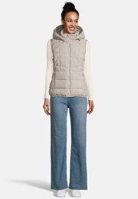 Lichtgrijze puffervest met een capuchon, met doorgestikte delen en een voorrits. Draag het over een crèmekleurig shirt met lange mouwen en in combinatie met blauwe jeans.