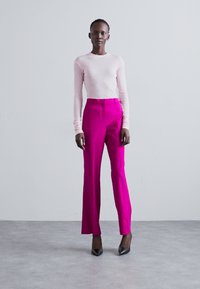 Vartalonmyötäinen vaaleanpunainen neulepaita yhdistettynä korkeavyötäröisiin kirkkaanpunaisiin housuihin. Mallilla on päällään mustat teräväkärkiset korkokengät. Sileä kangas ja muotoiltu design.