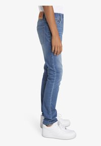 Jeans de denim azul ajustados con una cintura de altura media, corte recto y un ligero desgastado, combinados con zapatillas blancas. Parche de cuero marrón en la parte trasera.