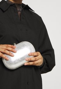 Osoba w czarnej koszuli trzyma małą, srebrną torebkę o teksturowanej powierzchni z zaokrąglonymi krawędziami i zapięciem na zatrzask.