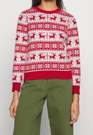 Donna che indossa un maglione natalizio rosso e bianco con motivi di renne e fiocchi di neve, abbinato a pantaloni verdi.