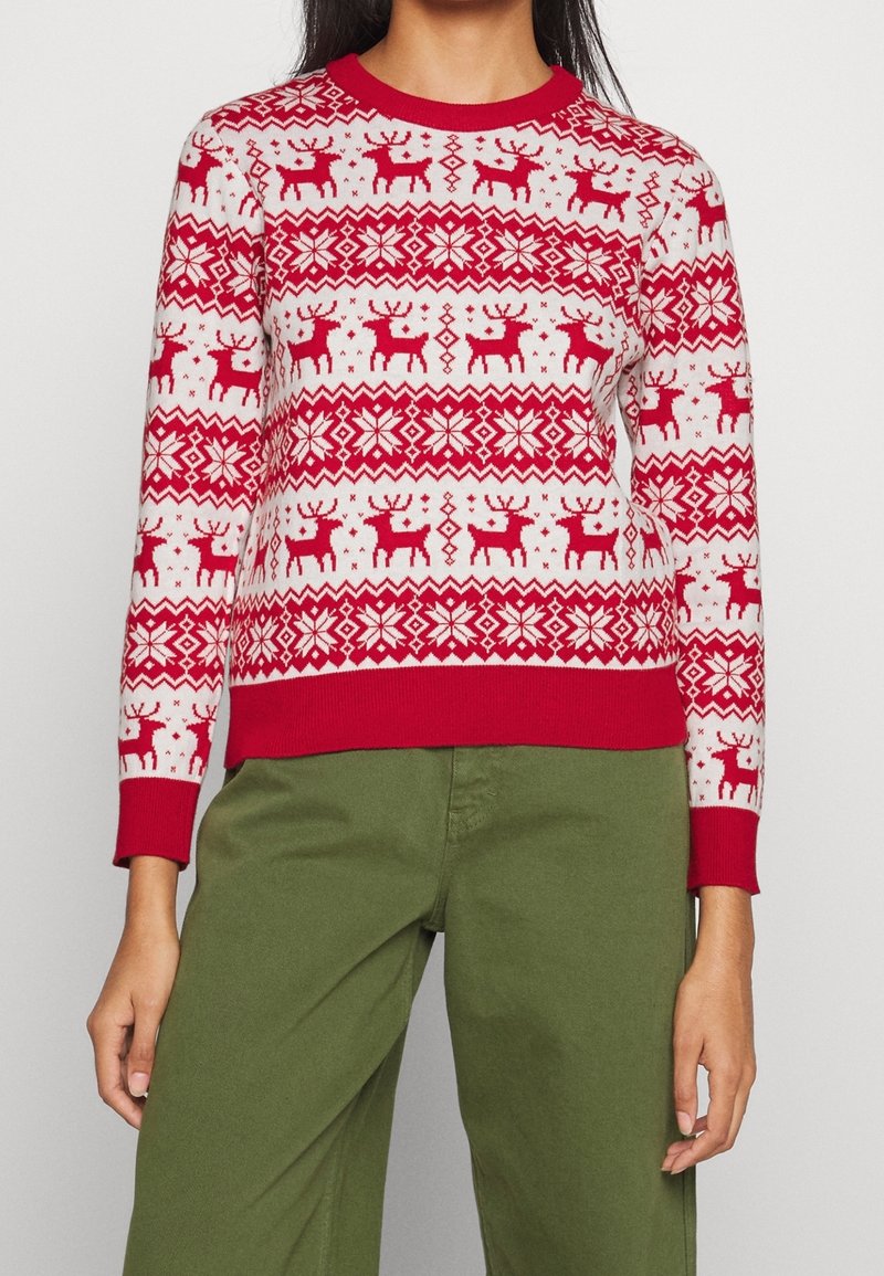 Donna che indossa un maglione natalizio rosso e bianco con motivi di renne e fiocchi di neve, abbinato a pantaloni verdi.