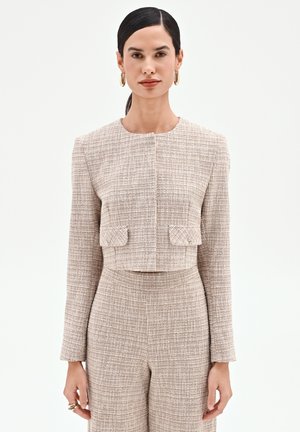 Femme portant une veste courte texturée beige et un pantalon taille haute assorti, debout avec les mains détendues le long du corps sur un fond uni.