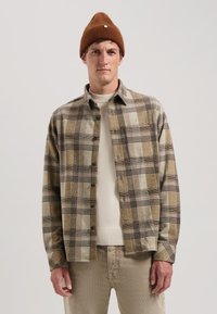 Cămașă în carouri maro și bej, cu guler standard, buzunar frontal și închidere cu nasturi, purtată peste un pulover crem și pantaloni bej, asortată cu o căciulă maro.