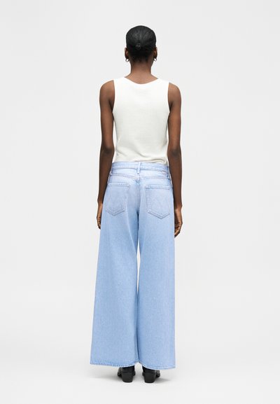 Lühikeste juustega inimene, kes kannab valget varrukateta topi ja helesiniseid laiade säärtega pükse, seisab seljaga vaataja poole, halli ükslabase tausta ees.