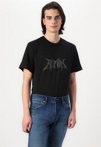 T-shirt en coton noir avec un logo graphique gris proéminent sur le devant, associé à un jean en denim bleu. Design simple à col rond.