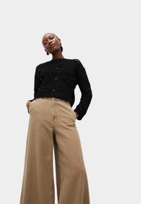 Pull en maille noir avec motif texturé et boutons, associé à un pantalon ample beige. Design simple, coupe décontractée et silhouette fluide.