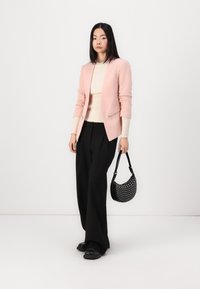 Light pink blazer with zippered pockets, beige ribbed sweater, black wide-leg trousers, and a black perforated handbag. Black shoes complete the look.
