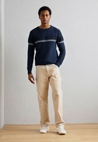 Marine stripete genser med teksturert finish, kombinert med beige bukser og hvite joggesko. Står mot en lys grå vegg på tregulv.
