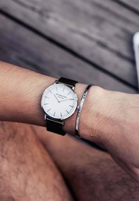 Orologio da polso in argento con quadrante bianco, cinturino in nylon nero e design minimalista. Accompagnato da un bracciale in argento con testo inciso.