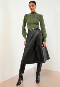 Grüner Kabelstrickpullover mit puffigen Ärmeln, kombiniert mit einem schwarzen, ledernen Midirock und kniehohen schwarzen Stilettos mit einem Gürtel.
