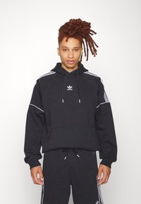 Felpa nera con cappuccio, dettagli bianchi e strisce sulle spalle, dotata di tasca frontale e cappuccio regolabile con cordino. Logo Adidas sul petto.