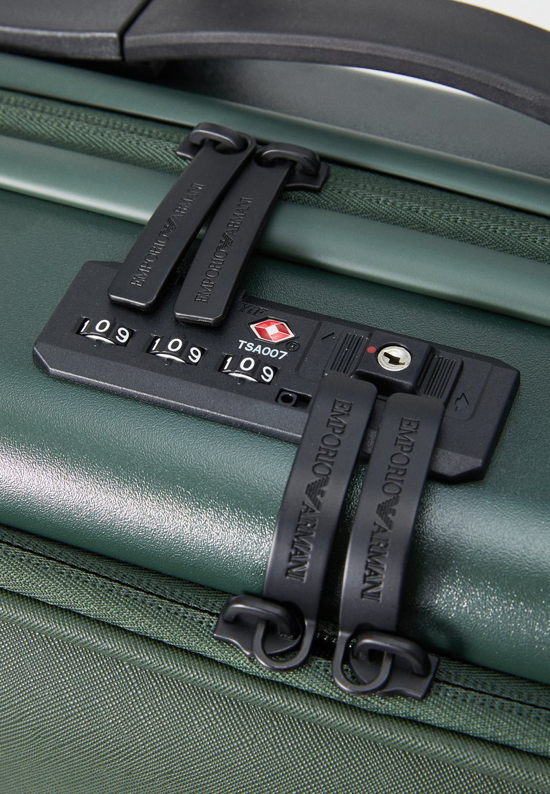 Maleta rígida verde con dobles cremalleras negras y candado aprobado por la TSA, que presenta una superficie texturizada y acentos de logo en relieve.