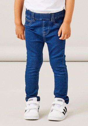 Jean en denim bleu avec une coupe slim, une texture lisse et des poches avant, associés à des sneakers blanches avec des rayures noires.