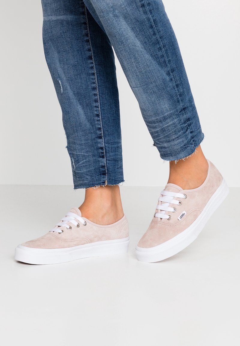 Persona che indossa sneaker Vans in suede rosa chiaro e jeans blu in denim a vita corta con orli sfilacciati su un pavimento bianco.