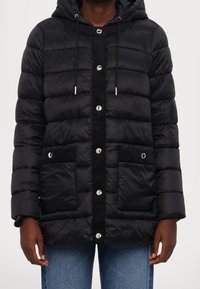 Chaqueta negra acolchada con capucha, botones a presión plateados, dos bolsillos frontales y un panel central texturizado. Forro de piel sintética en los bordes.