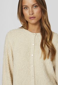 Cardigan color crema realizzato in un materiale testurizzato e soffice. Presenta un colletto rotondo e cinque bottoni decorativi lungo la parte anteriore.