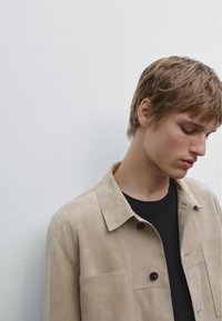 Beige suede jacket with a collar, front pockets, and black button accents, worn over a black shirt; shown against a plain background.