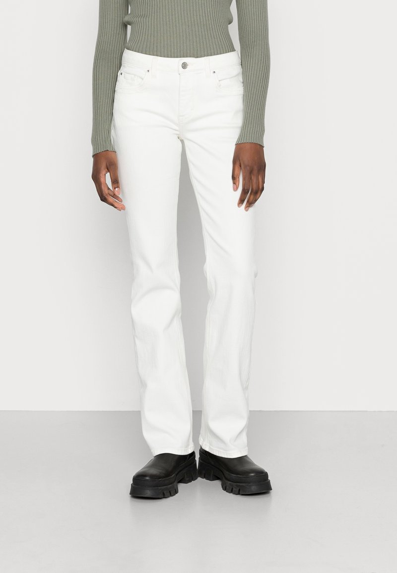 Personne portant un jean blanc droit, un haut côtelé kaki à manches longues et des bottes plateformes noires épaisses, debout devant un fond blanc.