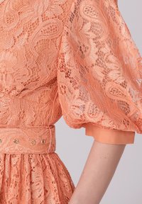 Robe en dentelle orange avec des manches bouffantes et une taille cintrée, ornée d'une texture en dentelle à motifs et d'une bande de couleur unie.