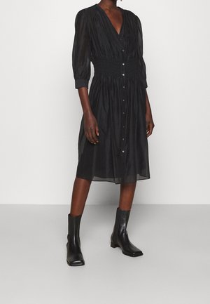 Personne portant une robe midi noire boutonnée à manches trois-quarts et des bottines noires, debout sur un sol gris devant un mur blanc.