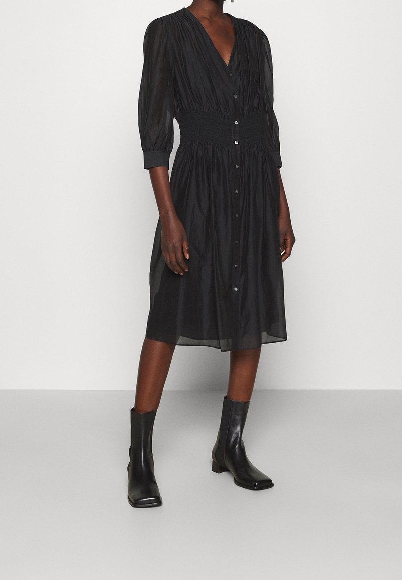 Personne portant une robe midi noire boutonnée à manches trois-quarts et des bottines noires, debout sur un sol gris devant un mur blanc.
