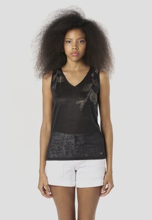 Črna brezrokavnik z V-izrezom, izdelan iz teksturirane tkanine z bleščečim cvetličnim okrasjem na ramenu. Kombiniran z belimi hlačnicami.