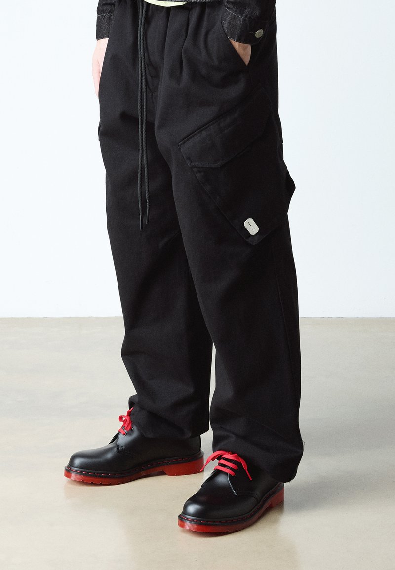 Personne portant un pantalon cargo noir ample et des chaussures en cuir noires avec des semelles rouges et des lacets rouges, debout sur un sol beige.