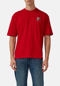 Rotes Baumwoll-T-Shirt mit Rundhalsausschnitt und kurzen Ärmeln, mit einem kleinen Snoopy-Graphic im linken Brustbereich. Getragen mit blauen Jeans.