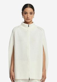 Vit stickad cape med hög krage, dragkedjeslutning och sidöppningar. Texturerad tyg med ribbade kanter, minimalistisk design.