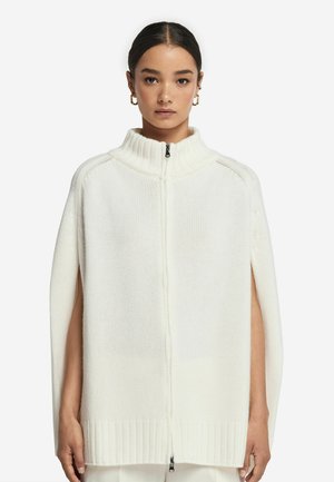 Hvid strikket kappe med høj krave, lynlås lukning og sidelukninger. Tekstureret stof med ribbede kanter, minimalistisk design.