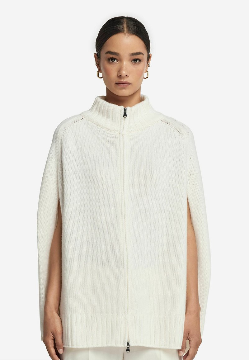 Vit stickad cape med hög krage, dragkedjeslutning och sidöppningar. Texturerad tyg med ribbade kanter, minimalistisk design.